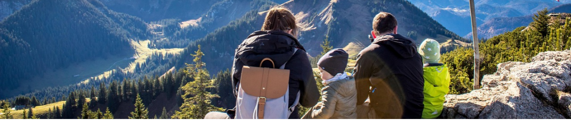 A family sitting together on a mountaintop, enjoying a scenic view of forested hills and valleys under a clear blue sky.